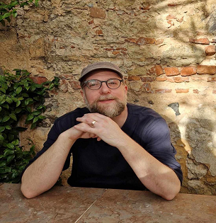A man with a beard, wearing black-rimmed glasses and a cap is smiling at the camera. He is sat down with his chin on his hands in front of a brick wall. 